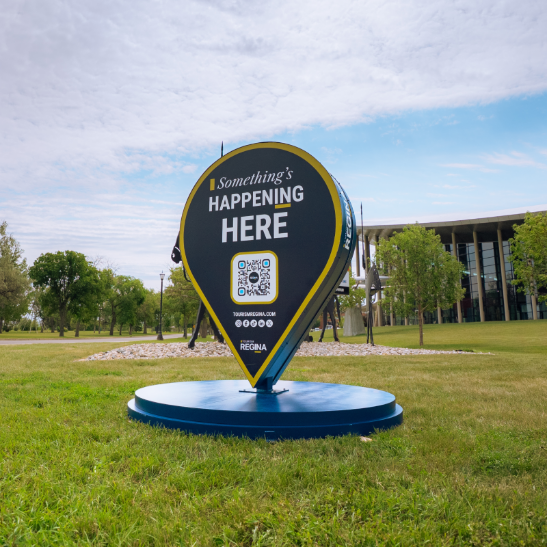 A Tourism Regina "Something's Happening Here" pin is placed on the lawn outside a Regina landmark inviting visitors to check out the attraction and scan the QR Code for more information. A Tourism Regina "Something's Happening Here" pin is placed on the lawn outside a Regina landmark inviting visitors to check out the attraction and scan the QR Code for more information.